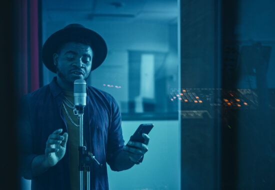 Portrait of Successful Young Black Artist, Singer, Performer Singing His Hit Song for the New Album. Wearing Stylish Hat, Holding Smartphone and Standing in Music Record Studio Soundproof Room.